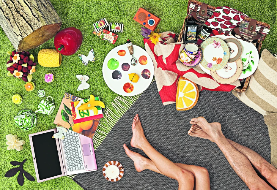 Overhead shot of picnic on green grass, blanket with food and drinks, open laptop, legs of a couple, still life, outdoor setting.