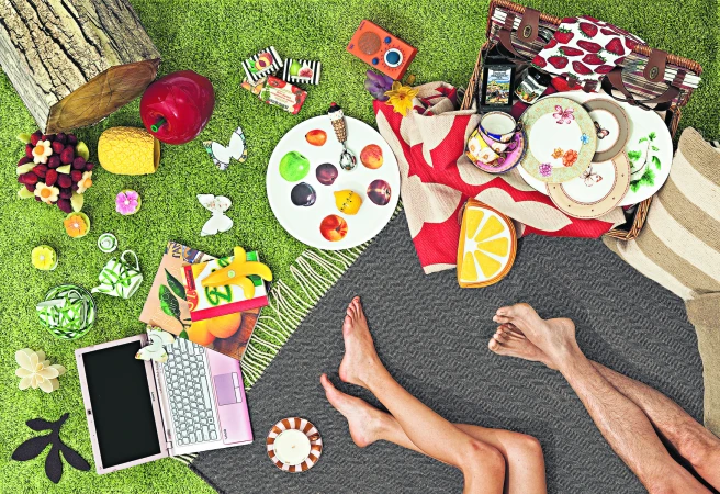 Overhead shot of picnic on green grass, blanket with food and drinks, open laptop, legs of a couple, still life, outdoor setting.