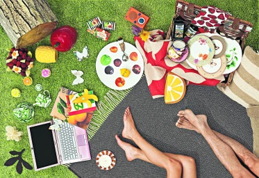 Top-down view of a picnic on artificial green grass with a basket, tableware, laptop, and bare legs.