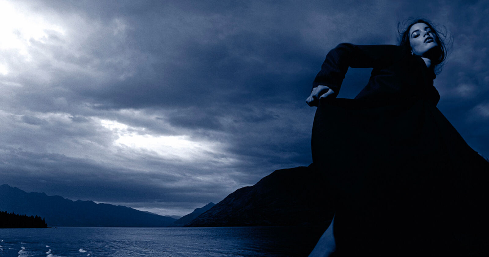 Woman in long black dress poses in front of lake and mountains with cloudy blue sky in outdoor fashion shot, low angle view.