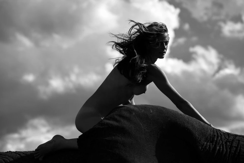 Striking black and white portrait of a bare female with windblown hair sitting atop an elephant against a cloudy sky backdrop.