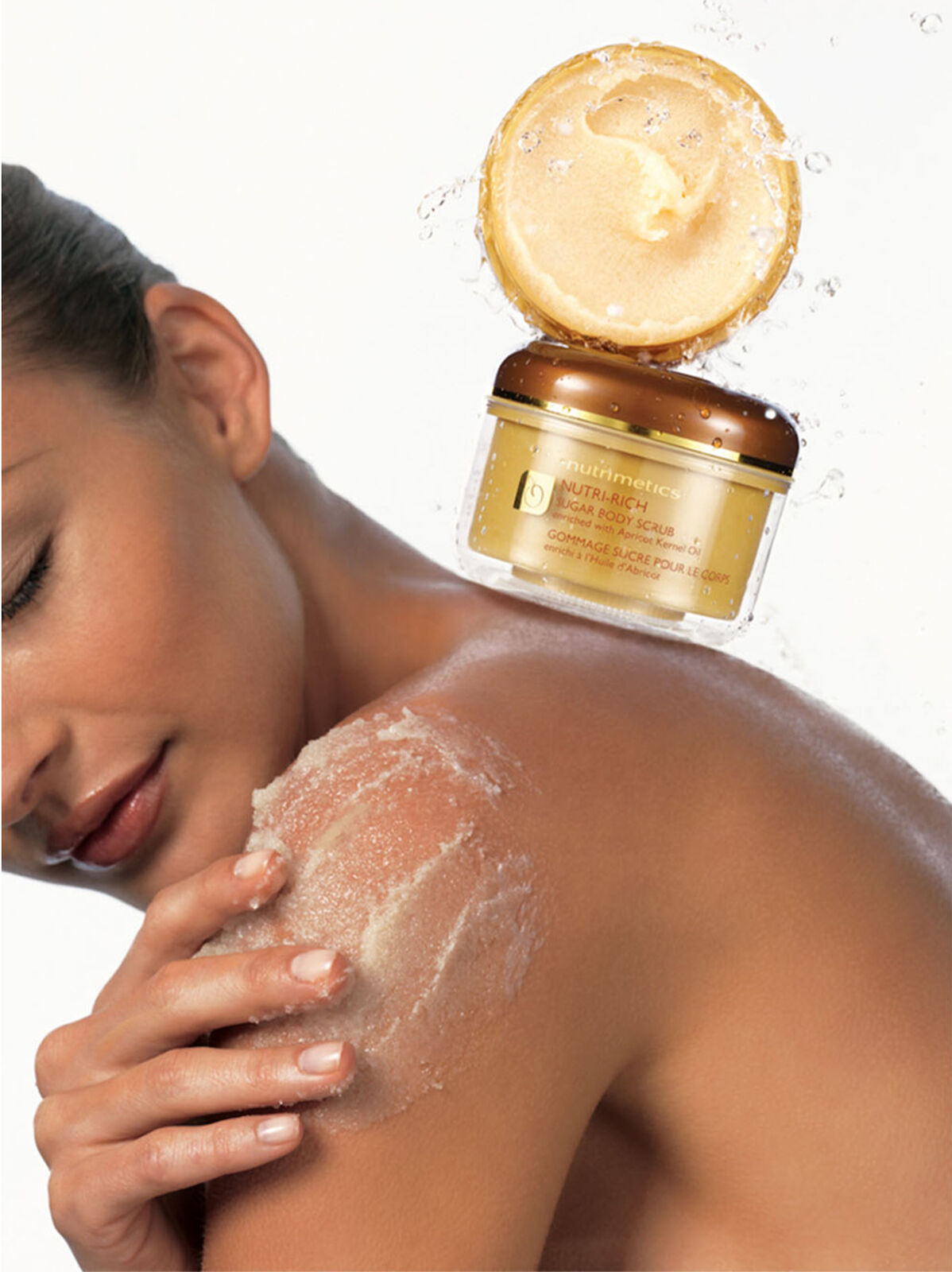 Woman applying sugar scrub to shoulder with product jar floating above in a beauty shot against a bright background with water droplets.