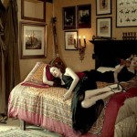 Two adult women lounge on an ornate bed in a vintage-styled indoor setting, surrounded by framed pictures and soft, diffused light.