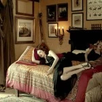 Two adult women lounge on an ornate bed in a vintage-styled indoor setting, surrounded by framed pictures and soft, diffused light.