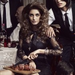 A woman in a lace dress sits at a table cutting meat with a knife, with other people in the blurred background and low lighting.