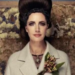 Adult female poses in a photo studio wearing a white overcoat with a floral brooch and a beaded headband, against a textured wall.