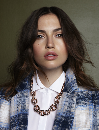 Close-up of a woman with dark hair and fair skin wearing a white shirt, blue plaid jacket, and gold chain necklace against green backdrop.