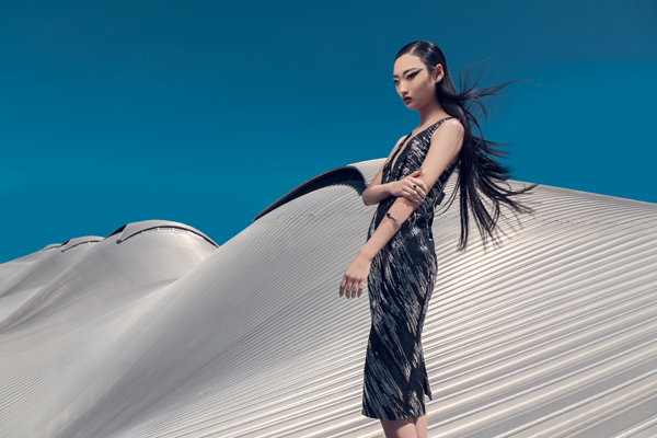 A slender Asian fashion model stands on an architectural surface against a clear turquoise sky, showcasing a metallic dress and sleek hair.