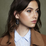 Close-up portrait of a woman with wavy brown hair wearing a tan coat and a light blue shirt against a neutral background.