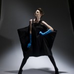 A female fashion model strikes a pose in a black dress, tights, and gloves against a gradient gray background, highlighted by studio lighting.