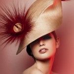 Elegant woman poses in a photo studio wearing an ornate beige couture hat with a red feather embellishment against a coral pink background.