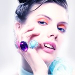 Close-up portrait of a fair-skinned woman with colorful makeup, wearing large gemstone rings, posing against a blurred white background.