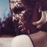 Portrait of a woman with a net veil over her face, featuring blonde hair and a blurred, natural background with soft, diffused lighting.