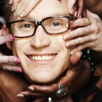 Close-up of a white adult male's face surrounded by the hands and arms of a group of diverse people touching his face.