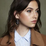 Portrait of a woman wearing statement earrings, light blue shirt, and brown jacket against a textured background