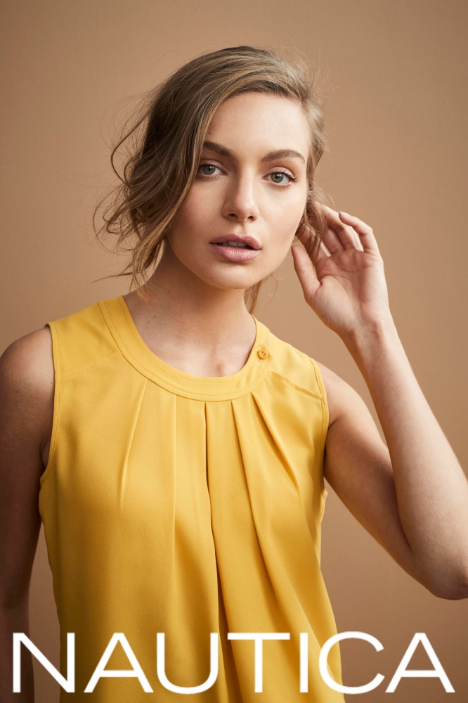 Close-up of a young adult woman in a yellow sleeveless top, posing against a tan background. The brand 'Nautica' is visible.