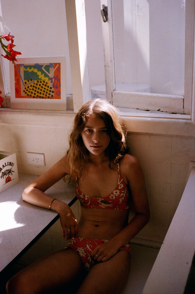 A young woman in a red floral bikini sits indoors near a window with sunlight, a painting, and a box with the word MALIBU.