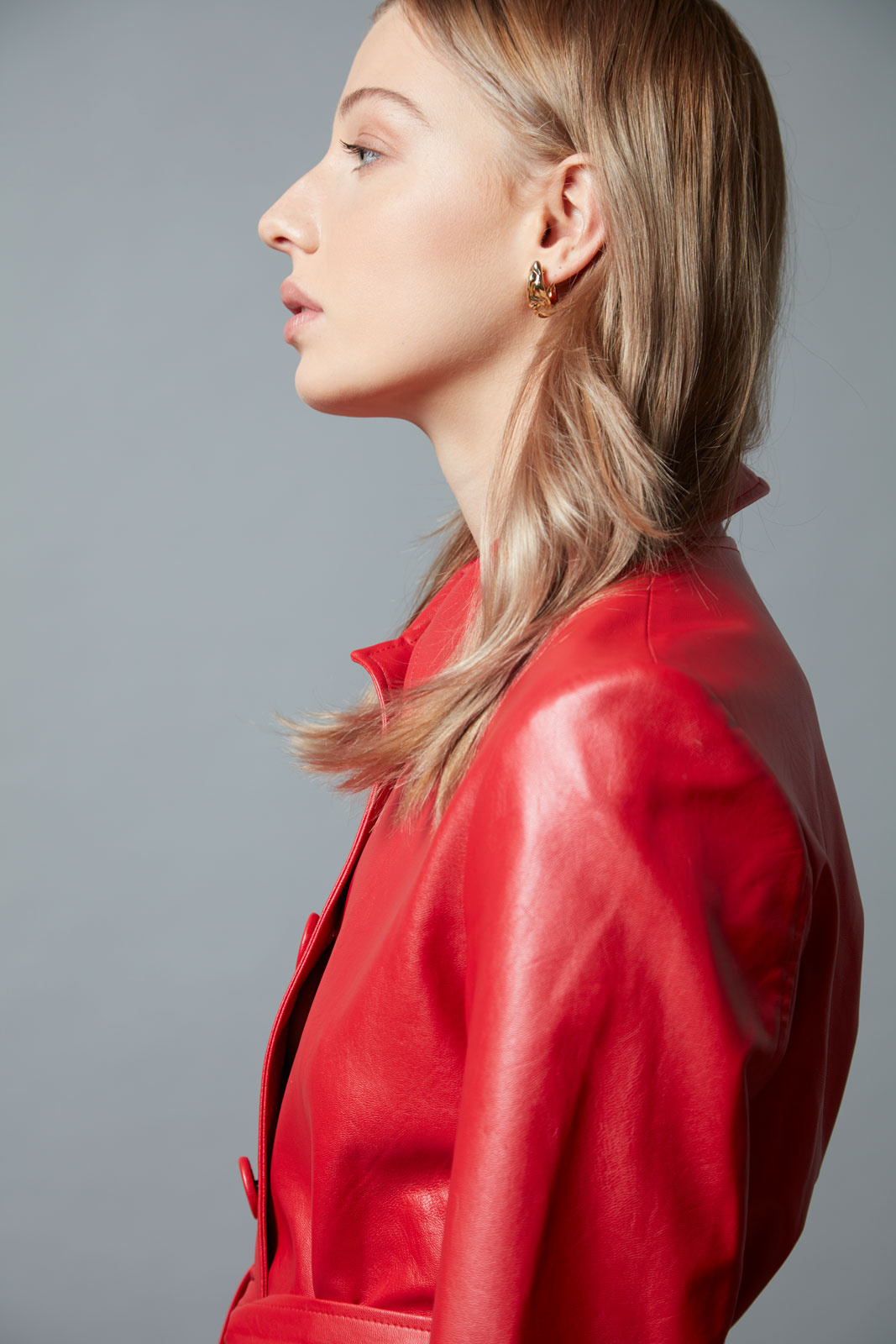 Profile shot of a woman with blonde hair wearing a bright red leather jacket against a light gray studio backdrop, looking off-camera.