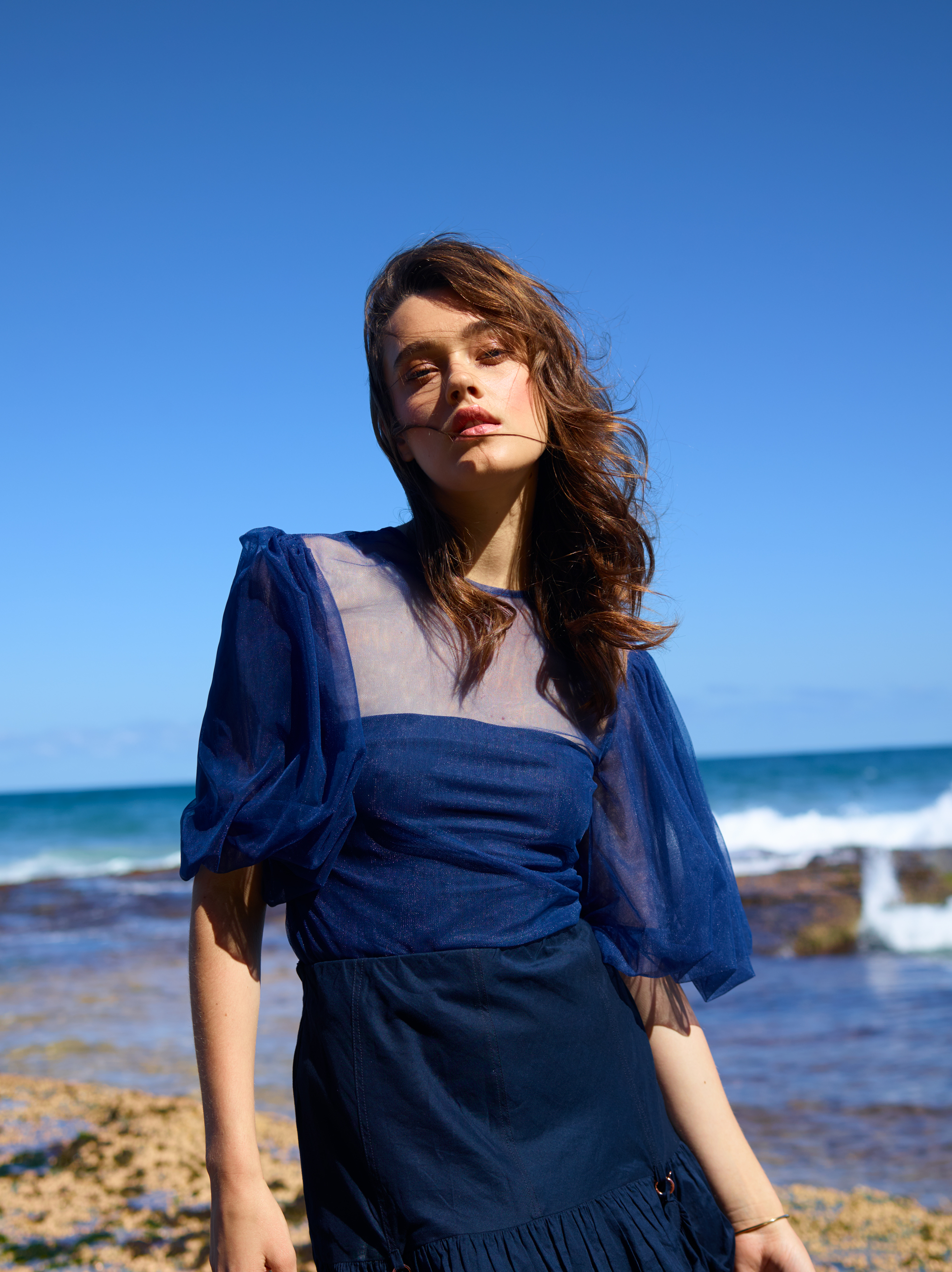 A young woman poses on a sandy beach with ocean waves crashing behind her, set against a clear blue sky, in a fashion editorial shot.