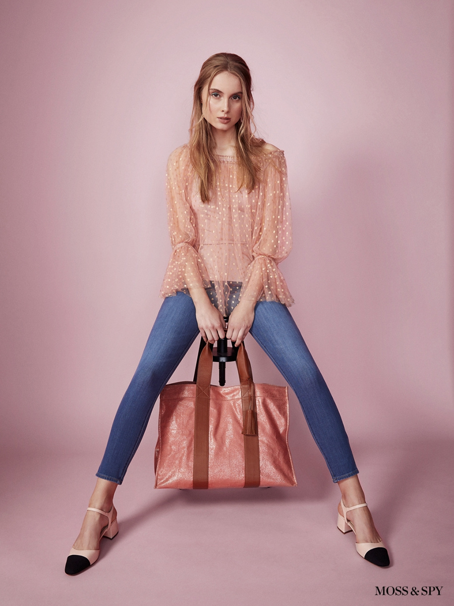 A fair-skinned fashion model poses with a pink metallic tote bag wearing a peach blouse, blue jeans, and black and white heels against a pink background.