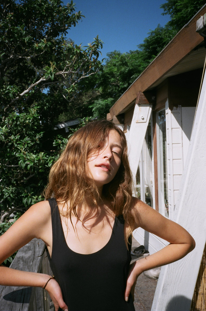 Outdoor portrait of a young woman in a black top with hands on her hips, standing on a wood deck near a house and lush trees.