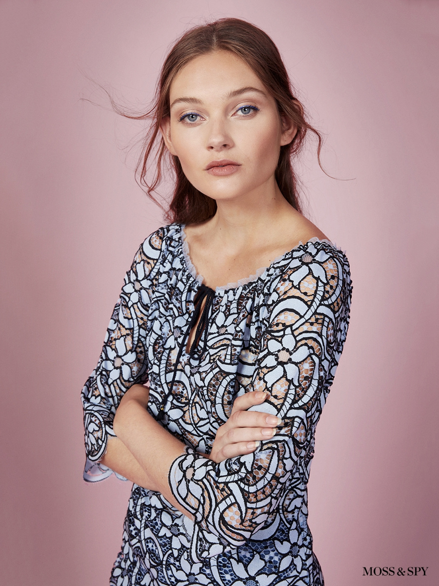 Close-up studio portrait of an adult female fashion model with long brown hair, fair skin, wearing a patterned blouse in front of pink backdrop.