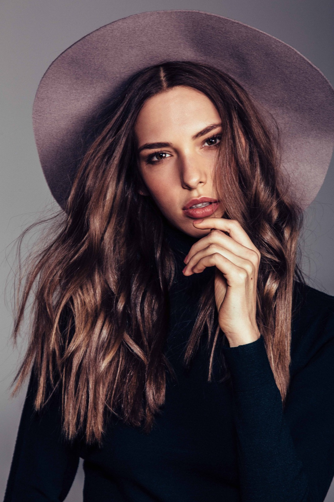 Portrait of a young adult female with long, wavy brunette hair and a wide-brimmed hat, posing with her hand near her face in a photo studio.