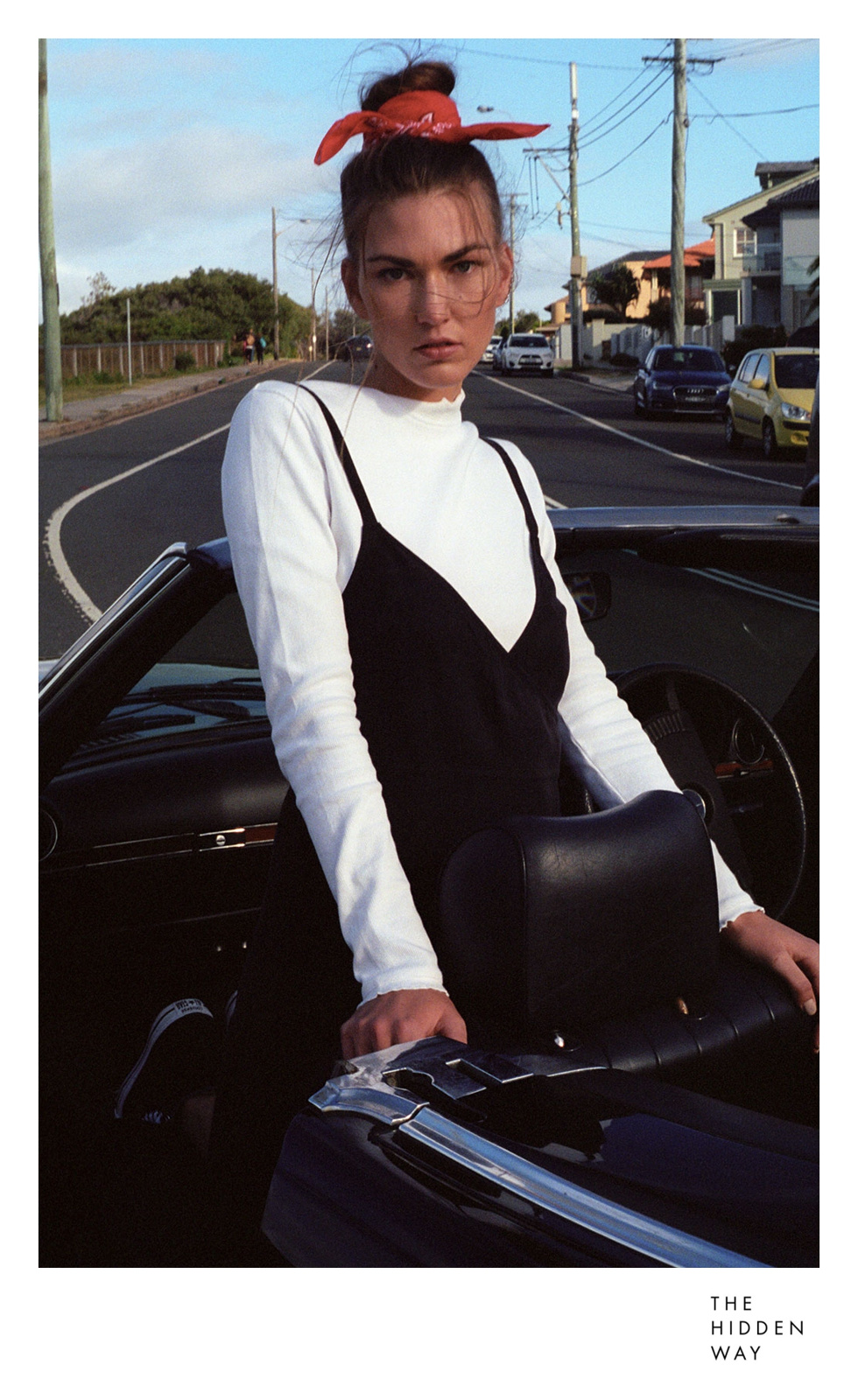 Adult female in a black dress, white long sleeve, and a red bandana standing next to a black convertible on an asphalt road.