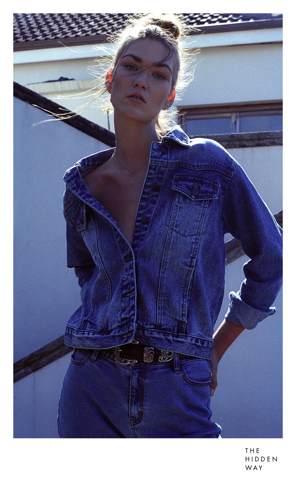 A woman poses outdoors wearing a denim jacket and jeans with a decorative belt. The photo has a shallow depth of field.