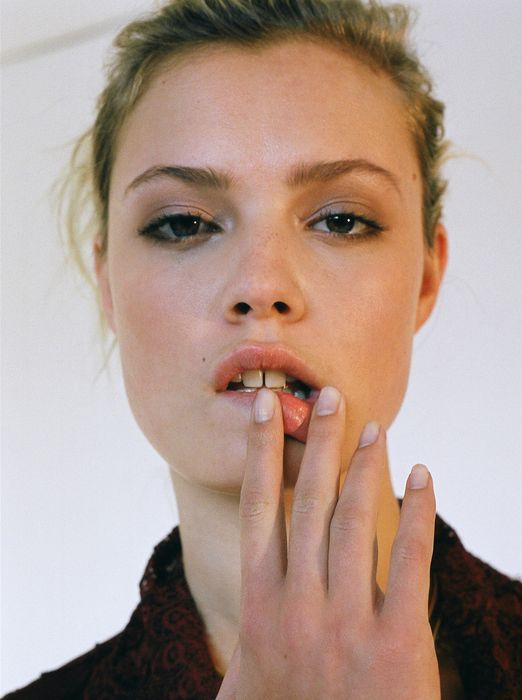 Close-up portrait of a fair-skinned young woman with blonde hair, brown eyes, and fair complexion, posing with hand near mouth against white background.