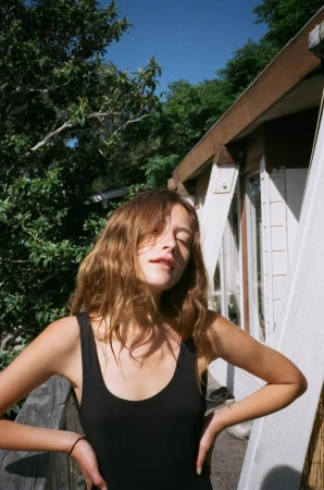 Young woman in a black tank top standing outdoors in bright sunlight near a building exterior.