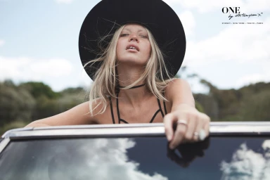 Blonde model in wide-brim black hat leaning out of a car window under an open sky, styled by Jessica Diez.