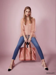 Model seated on a stool wearing a sheer dotted pink top and blue jeans, holding a large metallic pink tote bag against a pink background.