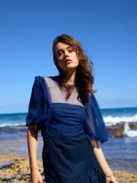 Model in dark blue mesh puff-sleeve top standing against a rocky coastal shore with ocean and clear blue sky.