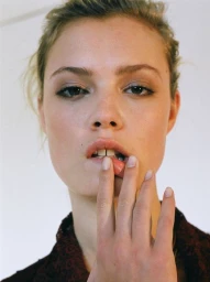 Close-up portrait of a woman with freckles resting her finger near her lips against a plain light background