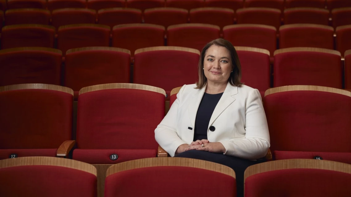 Portrait of a smiling woman with folded hands in a white blazer sitting in a red theatre seat with rows of empty seats behind her.