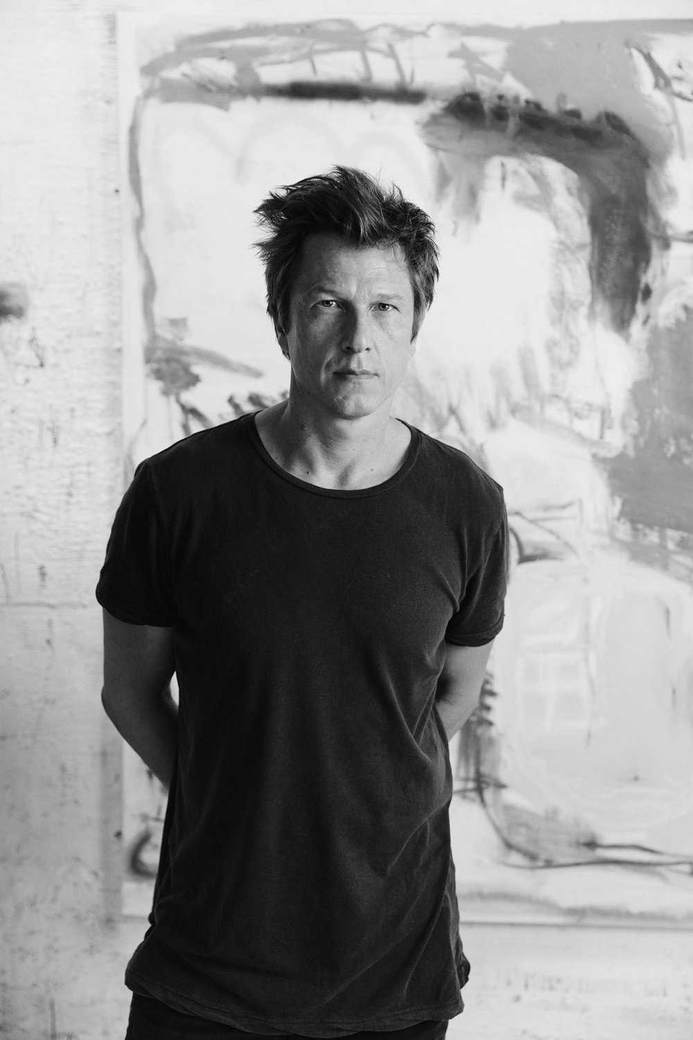 Black and white portrait of a man in a black t-shirt standing against an expressive abstract painted backdrop