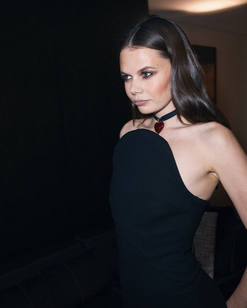 Portrait of a young woman in a black dress and statement necklace looking away against a dark background in an indoor setting.
