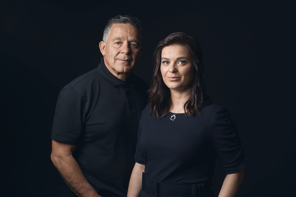 Portrait of an adult man and adult woman posing indoors in front of a dark background, wearing dark clothing with a medium studio shot.