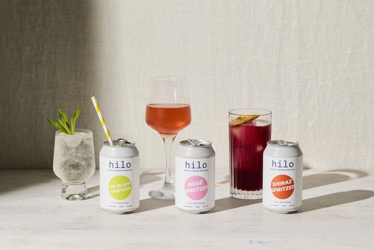 Canned spritzer drinks arranged with stemmed glasses and garnishes in a studio setting with soft, diffused light.