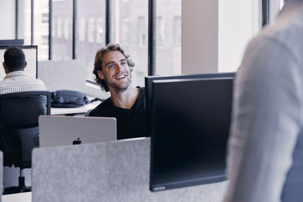 Man smiles in office setting, working at a desk with a laptop and computer monitor. Another person walks by in the foreground.