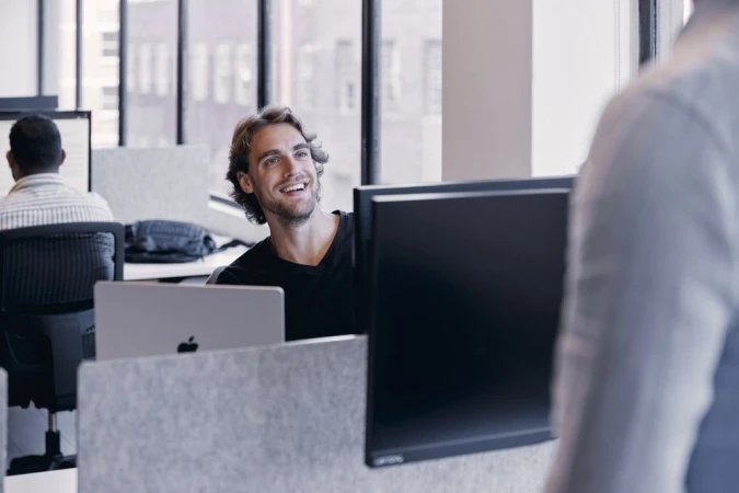 Man smiles in office setting, working at a desk with a laptop and computer monitor. Another person walks by in the foreground.