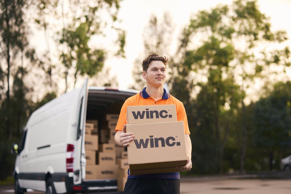 Delivery worker in uniform carries boxes with the winc. logo near a delivery van and a backdrop of trees under a sky.