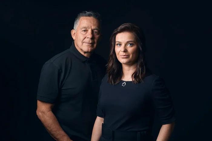 Portrait of an older man and a woman in black polo shirts standing against a dark seamless background