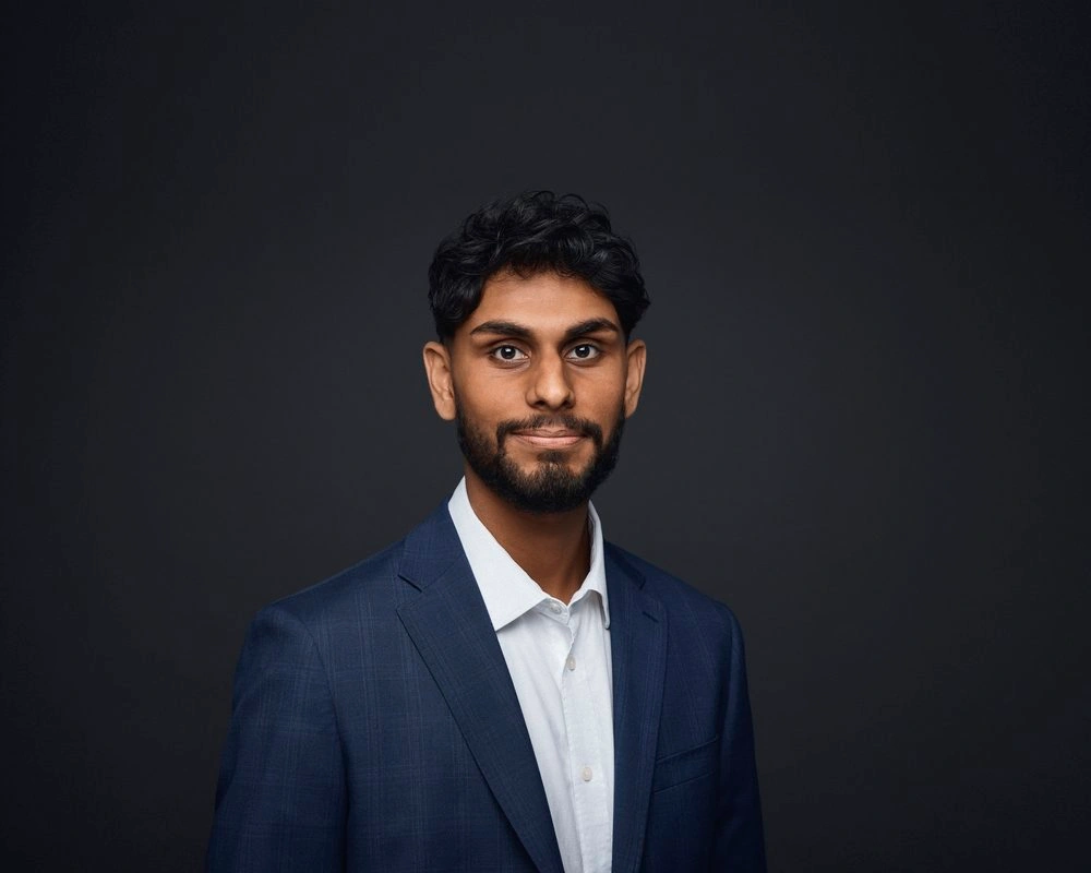 Portrait of a south asian man in a blue suit jacket and white collared shirt against a dark grey background.