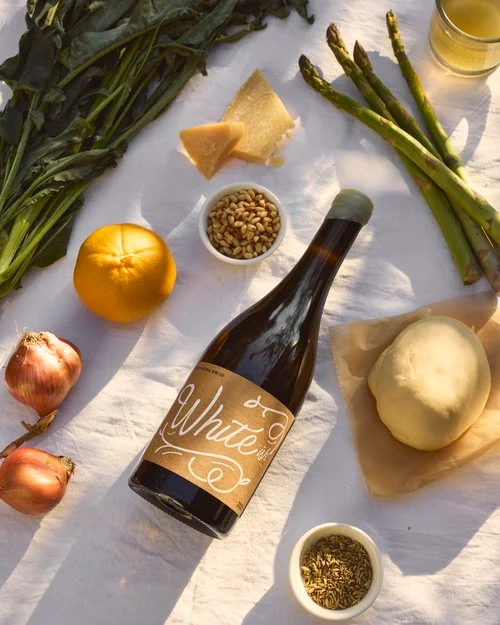 Overhead flat lay of a glass bottle with a white label, fresh asparagus, cheese, pine nuts, and vegetables on a white linen cloth.