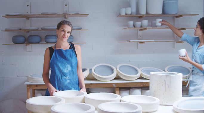 Two women work in a ceramics studio with shelves of pottery in the background. The first woman is wearing a blue apron.