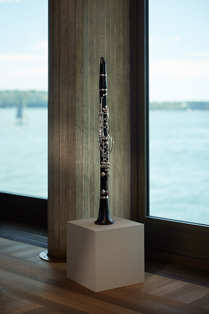 Clarinet stands on a podium in front of a window with a view of a lake and sky. The black clarinet is sleek and modern.