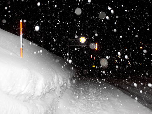 Snow-covered landscape at night with falling snow. A white pole and road traffic signals are visible in the background.