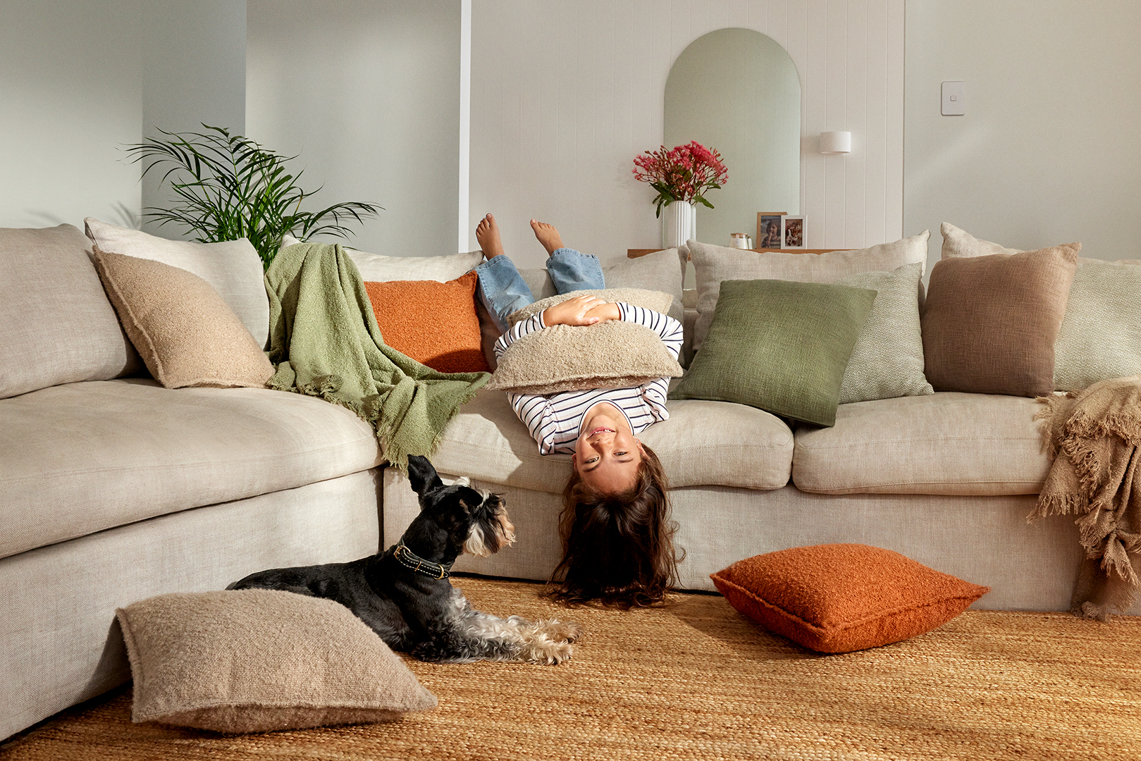 Young girl playfully hangs upside down on a cream-colored couch with her dog nearby in a warmly lit living room setting.